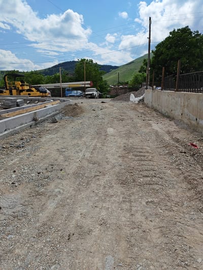 The image depicts an outdoor construction site on a sunny day. The foreground is a dirt and gravel road with tire tracks, suggesting recent vehicle activity. To the left, a yellow SAKAI SW 850 roller is parked near a newly constructed concrete curb and wooden forms. In the mid-ground, several vehicles are visible including a blue car, a white Lada Niva, and other construction equipment. Behind the vehicles, a chain-link fence and some buildings can be seen, with a pile of white material, possibly construction debris or a tarped load, nearby. The background features lush green hills and mountains under a bright blue sky with scattered white clouds. A utility pole with wires stands to the right, and a concrete wall runs along the right edge of the frame. The scene appears to be in a rural or semi-rural area, consistent with the provided location context of Bazum, Armenia. The overall impression is of ongoing infrastructure work in a scenic natural setting. There is some visible text on the roller: "SAKAI" and "SW 850". The white car has a license plate that appears to read "BL AC 964". There is also some text on a yellow piece of equipment behind the roller, possibly "22ANG".