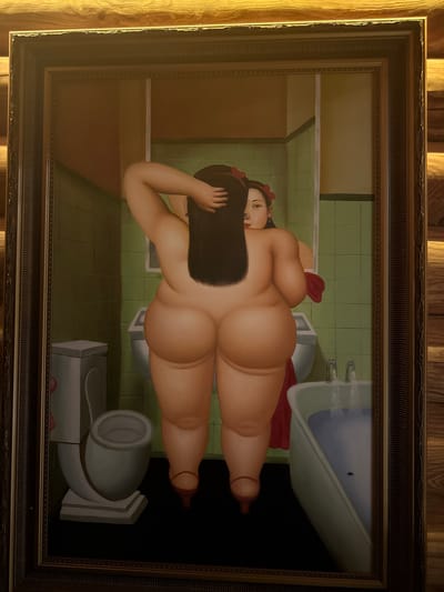 This is a painting by Fernando Botero, depicting a voluptuous nude woman lying in a bathroom. The woman is positioned in the center of the frame, with her back to the viewer, her head turned to the side to look at the audience. Her dark hair cascades down her back, and she wears a red ribbon in her hair. Her body is depicted in Botero's signature style, characterized by exaggerated volume and roundness.
The bathroom setting includes a bathtub filled with blue water, a toilet, and a sink. The walls are tiled in a pale green color. The floor appears to be made of large, rectangular tiles. The overall lighting suggests an indoor setting, possibly daytime, with no specific indications of time of day or weather. The mood of the painting is somewhat ambiguous; while the subject is nude and in a private setting, her gaze towards the viewer creates a sense of direct engagement. There is no visible text in the painting. The wooden frame surrounding the artwork suggests it is a displayed piece, possibly in a home or gallery. The external wooden paneling implies the painting is hanging on a wooden wall.