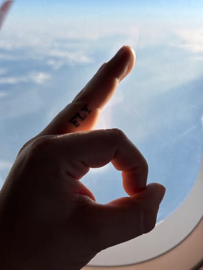 A hand is positioned against a window, creating a circle with the thumb and forefinger. The middle, ring, and pinky fingers are curled into the palm. The index finger features a tattoo of the word "FLY". The backdrop visible through the window is a blue sky with clouds. The image appears to be taken inside a vehicle, potentially an airplane, given the clear view of the sky. The location is not discernible from the image alone and the city Berg, Luxembourg is not visible.