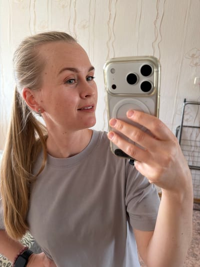 A fair-skinned woman with blonde hair pulled into a ponytail and a grey t-shirt is taking a selfie with her iPhone in what appears to be a room in Кульдур, Russia. She is wearing small hoop earrings and a black fitness tracker on her left wrist. The background shows a wall with vertical, wavy patterns.