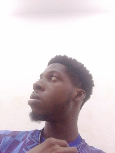 The media file is a close-up, waist-up portrait of a young Black man looking upwards and to the left. He has dark skin, short, curly black hair, and a goatee. He is wearing a purple and blue patterned shirt. The background is a plain, light-colored wall, possibly with some texture. The lighting appears to be natural and soft, suggesting it might be daytime. The overall mood is contemplative or perhaps aspirational due to his upward gaze. No text is visible in the image. The location is identified as Abeere, Nigeria.