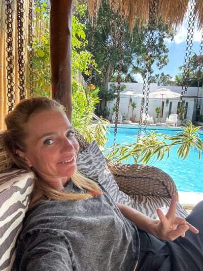 The image displays an adult female seated in a rattan-style swinging chair within an outdoor setting in Panglao, Philippines. She has light-colored hair and eyes, and a visible smile directed towards the camera. She wears a grey, possibly acid-wash or distressed, short-sleeved t-shirt and dark pants. Her left hand is raised, forming a V-sign with her index and middle fingers. The chair is suspended by chains from a structure featuring a thatched roof and a thick wooden support post. In the background, a bright blue swimming pool is visible. Beyond the pool, a white wall runs horizontally, fronted by several white lounge chairs and two large white patio umbrellas. Various potted plants and natural foliage, including trees and variegated plants, populate the area around the pool and extend into the distant background. The scene is brightly lit by natural sunlight, indicating a clear sky.