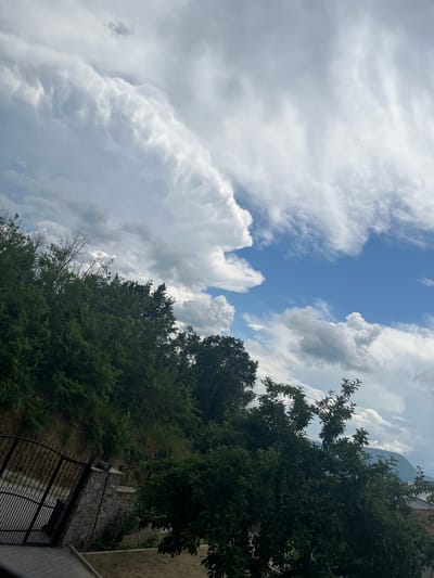 The image captures a sky with dramatic, billowing white clouds against a bright blue sky, with some gray storm clouds visible on the left. In the foreground, a lush green hillside is covered with dense trees. To the left, a black metal gate stands open, attached to a stone wall. The scene appears to be a natural landscape, possibly a park or a residential area on a hillside, in Ognyanovo, Bulgaria. There are no people or animals visible in the image.