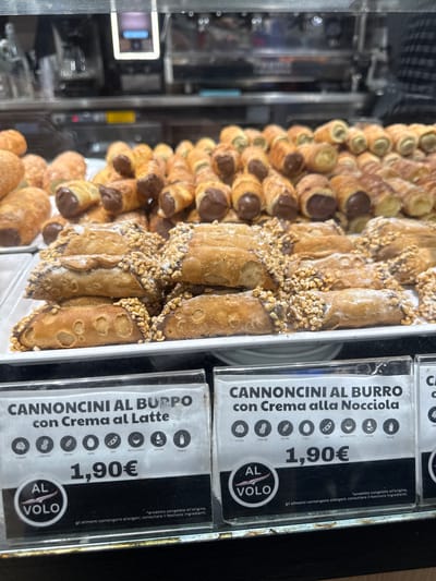 This is a close-up, eye-level, indoor shot of a display case at a food establishment, likely an airport or train station in Orio al Serio, Italy, given the "AL VOLO" branding which translates to "on the go." The foreground features two labels for pastries. The left label reads "CANNONCINI AL BURRO con Crema al Latte" and shows icons indicating potential allergens. Below this, the price is listed as "1,90€." The right label is for "CANNONCINI AL BURRO con Crema alla Nocciola" and also has allergen icons and the price "1,90€." The pastries themselves, cannoli-like tubes filled with cream, are visible in the background, some dusted with powdered sugar and nuts, and others with chocolate filling. The overall environment appears to be a food service counter with a blurred background of kitchen equipment.
