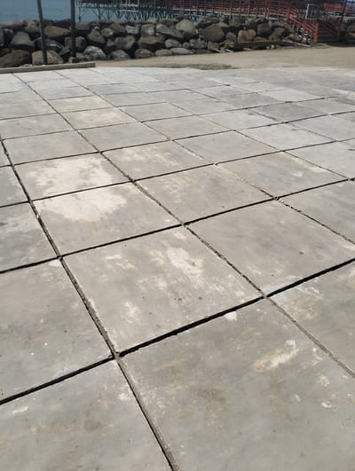 The image shows a paved area composed of large, square concrete tiles. The tiles are laid out in a grid pattern and exhibit signs of wear, with some discoloration and chips along the edges. The paving appears to be outdoors, possibly near a waterfront, as evidenced by the presence of a rocky embankment and a body of water in the background. Beyond the rocks, a pier or walkway structure with wooden elements and metal supports is visible. The scene suggests a coastal or harbor setting. The sky is bright, indicating daytime, and the lighting casts clear shadows, suggesting sunny weather. The overall impression is one of an open, public space with a utilitarian design. No people are visible in the image, and there is no discernable text. The location is identified as Lancok, Indonesia.