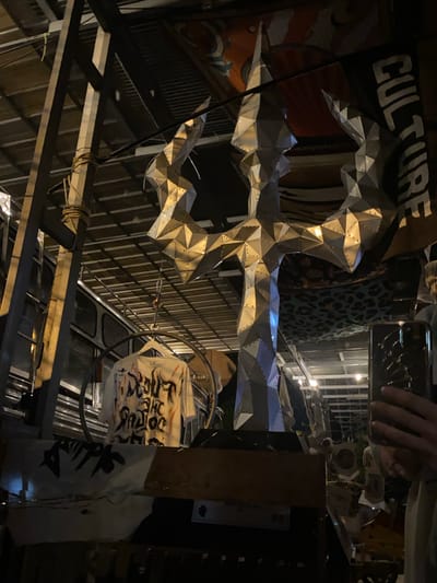 The image depicts an interior space with various objects and architectural elements. A large, silver, geometrically shaped object hangs prominently in the foreground. Behind it, a sign with the word "CULTURE" is partially visible. Other visible items include a rack, a printed bag, a metal cage, and various building materials and structures. A hand holding a mobile phone is visible in the top right corner, suggesting someone is taking a photo or video. Based on the prompt, this scene is located in Baan Nai Suan, Thailand.