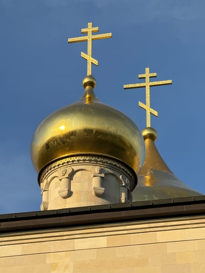The image shows the golden domes of the Resurrection Cathedral in Kirishi, Russia, against a clear blue sky. The foreground features a close-up of two of these domes, topped with Orthodox crosses. The larger, left dome is highly reflective, catching the sunlight and showing some signs of wear. The dome to its right is partially obscured and also bears the golden cross. Beneath the domes, decorative stone elements with sculpted faces are visible. The lower portion of the image reveals the light-colored stone facade of the building, with a dark metal trim. The scene is static, with no people or animals present, focusing solely on the architecture of the cathedral.