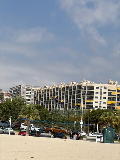 This media file captures an outdoor scene dominated by a large, multi-story building with numerous balconies, some adorned with yellow awnings. The building appears to be an apartment complex or hotel. In front of it, a net stretches across part of the frame, suggesting a recreational area like a volleyball court. Several vehicles, including cars and a pickup truck, are parked behind the net. Lush green trees and palm trees are interspersed throughout the scene, adding a tropical or Mediterranean feel. The ground in the foreground is a sandy or paved area. The sky above is partly cloudy. Visible text on one of the buildings reads "EDIFICIO CORUNA". The overall impression is a sunny day in a coastal urban or resort setting.