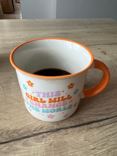 An orange-rimmed, white ceramic mug with the text "THIS GIRL WILL CHANGE THE WORLD" printed on it in colorful letters and flower graphics, sits on a light brown wooden table. The mug contains dark liquid, likely coffee, filling about a quarter of its volume. The background shows a blurry interior with light-colored walls and a doorway with bags. The scene is devoid of people. The image does not provide any information regarding its location in the city of Rumia, Poland.