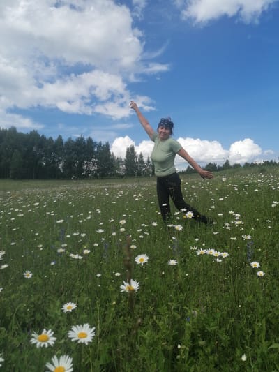 A woman is standing in a field of daisies under a blue sky with fluffy white clouds. She is wearing a green t-shirt, black pants, and a dark bandana. Her right arm is raised, and her left arm is extended to her side as if she is dancing or posing. The field is covered in numerous white daisies and green grass. In the background, a dense line of trees forms a dark green border. The scene appears to be set on a sunny day, likely in the late morning or early afternoon. The overall mood is cheerful and idyllic, suggesting a relaxed outdoor activity in a rural setting. The location is identified as Lagerevo, Russia. There is no visible text in the image.