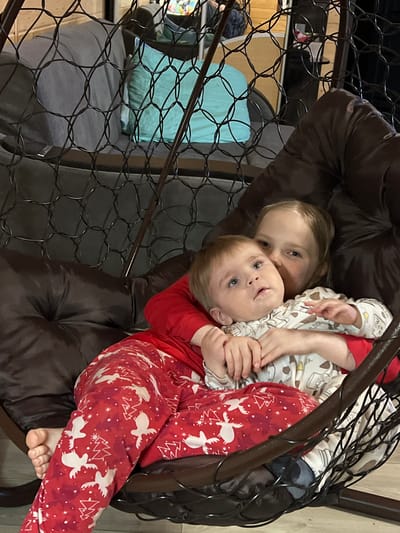Two Caucasian children, one older girl and a younger toddler, are sitting in a brown, cushioned hanging chair within a room. The toddler is wearing red and white holiday-themed pajamas and is cradled in the girl's arms. The girl is wearing pajamas with a cream-colored base and a cartoon animal pattern. The chair has a dark metal frame and a wire-like backing. In the background, another chair is visible, along with what appears to be a light blue pillow. The exact location within the city of Bol'shakino, Russia, is indeterminable.