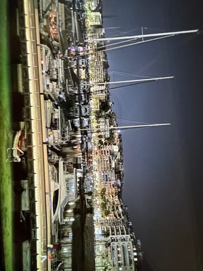 The image depicts a nighttime scene of a marina or harbor in the city of Senokos, Bulgaria. Numerous vessels are docked along a paved quay, which occupies the left side of the frame. The water, calm and dark, reflects ambient light. Prominently visible are multiple boats, including several large sailboats distinguished by their tall, slender masts extending upwards into the dark night sky. Other docked vessels appear to be motor yachts of various sizes.
In the background, a dense urban landscape rises, consisting of multi-story buildings that are brightly illuminated, creating a distinct skyline along the waterfront and extending up a gentle incline. The artificial lights from the buildings cast a glow over the scene. Scattered green vegetation, possibly palm trees, is discernible among the buildings. The sky above is dark and uniform, indicative of nighttime conditions with no visible celestial bodies. On the quay in the foreground, a small, light-colored dinghy with red accents is visible. No discernible individuals are engaged in specific actions, presenting a static scene focused on the illuminated urban and maritime infrastructure.