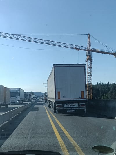 This media file captures a scene of heavy truck traffic on a highway in Hrusice, Czechia. The primary subjects are numerous semi-trailer trucks lined up on the road, indicating a traffic jam. The perspective is from inside a vehicle, looking forward through the windshield.
In the background, a large construction crane with the "STRABAG" logo is prominent against a clear blue sky. The sun appears to be high, suggesting it is daytime, likely mid-morning or afternoon. The weather seems fair with no visible precipitation. The highway is paved with clear yellow lane markings, and a concrete barrier separates lanes in some areas. A dense forest is visible on the right side of the road, beyond the barrier.
Visible text includes "STRABAG" on the crane and "1BD 6456" and "SCHMITZ" on the license plate and rear of the leading truck, respectively. The overall mood is one of slow progress and congestion due to the traffic. The scene is likely a common occurrence on major roadways, especially near construction sites.