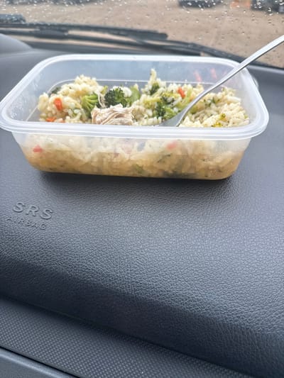A clear rectangular plastic food container, partially filled with a meal of rice, broccoli florets, shredded light meat, and small red vegetable pieces, rests on a dark, textured car dashboard. A silver-colored fork is embedded in the food on the right side of the container. The dashboard bears the embossed text "SRS AIRBAG" in its lower-left section. Beyond the windshield, which is speckled with raindrops, an out-of-focus background indicates an outdoor setting. The scene is observed within a vehicle in the city of Dawley, United Kingdom. No individuals are present in the image.