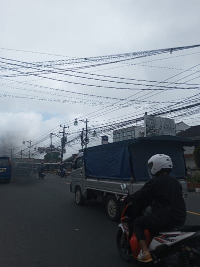 A street scene depicts a road with various vehicles, including a blue car emitting dark smoke, a small truck with a blue tarp, and a red and white motorcycle with a rider in a white helmet. Power lines crisscross the sky above street lights and signage. The sky is overcast, creating a muted atmosphere.