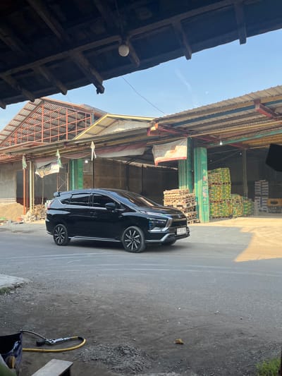 A black car is parked in Paweden, Indonesia, adjacent to industrial buildings, one stacked with goods and pallets. A weathered wooden structure is visible on the left, and the sky is partially visible. A hose and cleaning equipment are nearby.