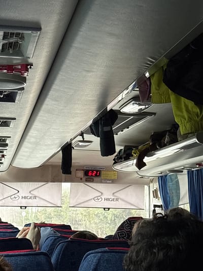 The image, taken inside a bus, shows an interior view with overhead storage, seats, and windows. The bus has a digital display showing "6:21" and branding for "HIGER" on the window panel. Passengers occupy the seats. There are indications of a city environment visible through the windows, which suggests the bus is moving. Based on the image metadata or any explicit indication, the location can not be definitively confirmed as Челва, Russia.