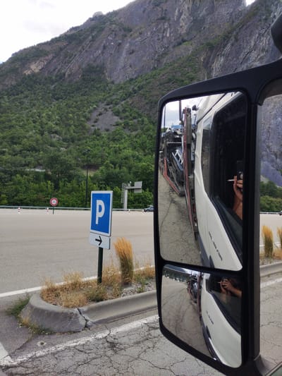 The image shows a reflection in the side mirror of a truck. The mirror shows a person holding a cigarette and a mobile phone in their hand, with a cigarette in their mouth. In the reflection, there are several cars loaded onto a car carrier truck. The background shows a steep, rocky mountain slope covered with trees and vegetation, with a cloudy sky above. A parking sign with an arrow pointing to the right is visible to the left of the truck. The scene appears to be in a mountainous area, possibly a rest stop or parking area for trucks. The location is identified as Saint-Martin-d'Arc, France.
