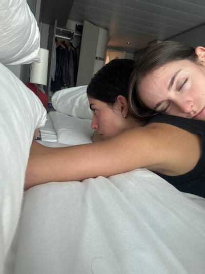 The scene is a cruise ship cabin, identified by its compact design, built-in furniture, and panelled ceiling, currently located at or in transit near San Miguel de Cozumel, Mexico. Two females are depicted lying in a bed with white bedding. The individual on the right is asleep, her eyes closed, with her head resting on the upper back of the individual on the left. She is wearing a dark sleeveless garment. The individual on the left is awake, positioned with her profile facing towards the left side of the frame. She has dark hair pulled back and a small hoop earring in her visible ear. Her right arm extends forward onto the white sheet. In the background, to the left, a white lampshade with a red base is visible next to an open closet containing dark clothing on hangers. A single dark hair strand is present on the white sheet in the foreground.