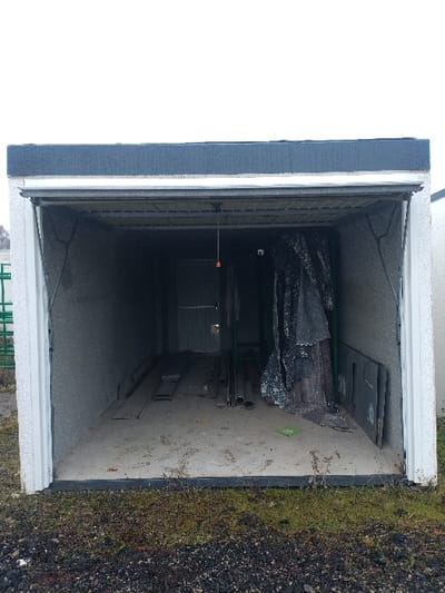 The image depicts an open, single-stall garage located in Bitburg, Germany. The garage exterior walls are light gray with a textured finish, topped by a dark gray flat roofline. The ground immediately in front of the garage consists of dark soil and gravel mixed with sparse patches of green and brown vegetation.

The overhead garage door is fully retracted, revealing the interior. The garage features light gray, textured concrete block walls and a light gray concrete floor, showing signs of dirt and wear. A dark gray overhead track system is visible for the garage door mechanism. A white pull cord with an orange handle hangs from the ceiling near the center of the garage entrance. No individuals are present, and no actions are depicted.

At the rear of the garage, a closed, dark-colored door, possibly metal, is visible. Along the left interior wall, several wooden boards and planks are arranged horizontally on the floor. In the mid-right section of the garage, a collection of cylindrical metal objects, likely pipes, rests horizontally. Further to the right, a large, dark-colored tarpaulin is draped over an unseen object, leaning against the right wall. Adjacent to the tarpaulin, two dark, flat rectangular panels lean vertically against the right wall. A small, bright green circular marker with a white numeral "0" is present on the concrete floor near the tarpaulin. High on the rear right wall, a small, white square object, consistent with a security camera, is mounted.