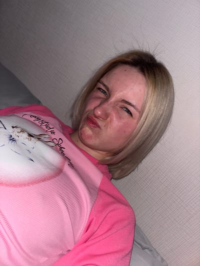 An indoor, close-up photograph features a young woman with shoulder-length blonde hair and darker roots, making a playful or exaggerated "disgusted" expression. Her eyes are squinted, nose wrinkled, and lips pursed. Her cheeks show a slight flush.
She is wearing a long-sleeved pink top. The sleeves and upper part of the shirt are made of a ribbed, textured fabric. The central panel of the shirt is a smoother, lighter pink, featuring a graphic print of a light-colored rabbit surrounded by delicate blue and purple floral designs. Above the rabbit, cursive text reads "cozystudio School," and below, partially visible text says "abbit princess."
The background consists of a light-colored wall with a subtle, textured geometric pattern, possibly wallpaper. To the right of the subject, a soft gray fabric, likely a pillow or bedding, is visible. The lighting suggests an indoor setting, likely artificial. The woman is angled, with the shot taken from a slightly low perspective. The scene is set in Ligolambi, Russia.
