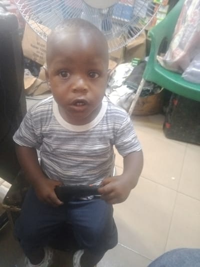 In an indoor retail or storage area within Akodo, Nigeria, a young, dark-skinned child, approximately 1-3 years old, is seated, facing the camera directly. The child has a shaved head and wears a short-sleeved t-shirt with horizontal white and grey stripes over dark blue trousers or shorts, paired with white and grey sneakers. The child's mouth is slightly ajar, revealing lower front teeth. Both hands hold a dark, elongated object, consistent with a remote control, horizontally across the lap. The background includes an oscillating fan with a metal cage positioned behind the child's head. To the left, a partially visible cardboard box displays "Mobil Super 1000" and "APL SN 20W-50." To the right, a green plastic chair, a wicker basket containing various items, and additional cluttered storage are discernible. The floor is surfaced with light-colored tiles.