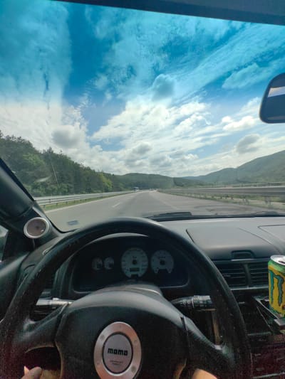 The image is a first-person perspective from inside a car, looking forward through the windshield. The car is on a highway, with guardrails lining the right side and a dense forest on a hillside to the left. The sky is blue with scattered white clouds. The dashboard of the car is visible, including the steering wheel, instrument cluster, and a can of Monster Energy drink. The steering wheel has a "momo" logo in the center. Based on the presence of the highway and the surrounding natural landscape, the scene is likely on a highway in or near Potop, Bulgaria.