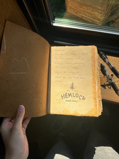 A first-person overhead view captures an open guestbook inside a cabin identified as Hemlock Hideaway, located in Otego, United States. The guestbook, with an aged, yellowish paper page on the right and a brown, suede-like cover on the left, is illuminated by direct sunlight.
On the right page, handwritten text in black ink reads: "09/27/2025 We are so happy to be the first guests in this amazing cabin. Celebrating our friend today! She is 24 today ♡ Rotem Mor Roy Niam Amie ♡♡♡". Below the text, a printed logo features a pine tree above "HEMLOCK HIDEAWAY EST. 2025". The page exhibits irregular brown staining, suggesting age or liquid contact.
A person's left hand, wearing a plain gold band on the fourth finger, holds the left page of the guestbook open. This left page, part of the inner cover, shows a faint white chalk-like outline drawing resembling a broad 'M' shape.
To the right of the guestbook, two black rollerball pens rest on a dark wooden surface. The upper pen is a standard design, while the lower pen features a complex metallic clip mechanism.
The background reveals a window with a dark frame, reflecting exterior elements including a wooden structure and green foliage. Below the table surface, the person's lap, clad in light blue-grey denim pants, and portions of dark footwear are visible.