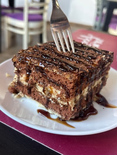 A close-up, slightly angled shot shows a slice of marbled chocolate cake on a white plate. The cake has layers of dark chocolate and lighter sponge, with a rich chocolate sauce drizzled over the top. A silver fork is positioned on the left, with its tines touching the cake. The plate is set on a dark surface, possibly a table, next to a burgundy placemat or tablecloth. In the background, out of focus, are elements of a cafe or restaurant setting, including what appears to be a pink paper item with some faint, elegant script, possibly a menu or a brand logo, and some blurred white furniture with purple accents. The lighting suggests it's daytime, and the overall mood is one of indulgence and enjoyment of a dessert. No specific emotional cues are evident from the image itself. The text in the background is too blurry to read. The location context is Chavdar, Bulgaria.