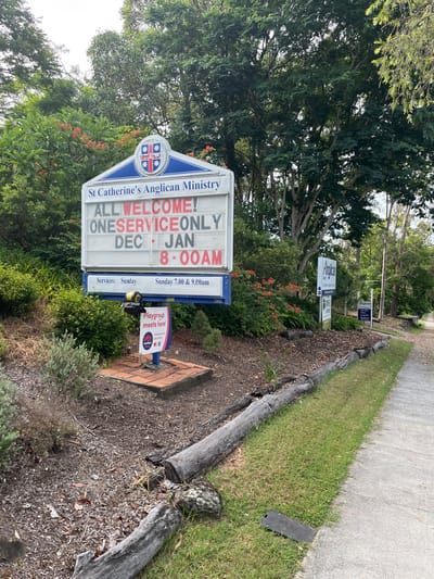 A daylight image captures a large sign for "St Catherine's Anglican Ministry" in what appears to be a community or church property in Riverhills, Australia. The sign, predominantly white with blue accents and a crest, stands prominently in the foreground. Visible text on the sign reads: "ALL WELCOME!", "ONESERVICEONLY DEC JAN 8-00AM", and below that, "Services: Sunday 7.00 & 9.00am". A smaller, colorful sign attached near the base of the main sign states "Playgroup meets here!".

The sign is situated next to a mulched garden bed, which features various green shrubs and some plants with small reddish flowers. A concrete sidewalk runs along the right side of the frame, bordered by a strip of green grass and a decorative edge made from stacked, weathered wooden logs. In the background, numerous large trees with lush green foliage create a natural, verdant backdrop. Another, smaller sign is partially visible further back among the trees, with the text "Aholica 43" discernible. The scene is illuminated by clear daylight, suggesting a bright, possibly overcast day. No people are visible, but the signs indicate scheduled church services and playgroup meetings as ongoing events at this location.