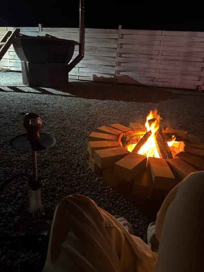 The scene depicts an outdoor setting at night. A fire is burning within a circular fire pit constructed of light-colored rectangular blocks. A wooden fence is situated to the left of the fire pit, and a black chair is positioned nearby. A hookah is placed on the ground next to the fire pit. The ground surface appears to be gravel. A person is partially visible in the top right, wearing light-colored clothing and shoes. The image is rotated counterclockwise. The location is not specified.
