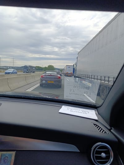 The image shows a traffic jam on a highway, likely the A3, near Marktheidenfeld, Germany. The perspective is from inside a vehicle, looking forward. Several large trucks and cars are visible in front. The sky is overcast with grey clouds. A white piece of paper with text is placed on the dashboard of the vehicle in the foreground. The text is in Czech, and it translates to "Permit to enter". The license plate of the car in front is visible and reads "PV-359-T". There are power lines and pylons in the background, indicating an industrial or roadside area. The overall scene depicts a common traffic situation on a major European highway.