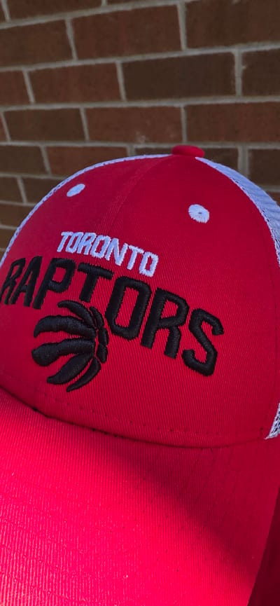 The image is a close-up, low-angle shot of a red baseball cap with "TORONTO RAPTORS" embroidered in white and black lettering, respectively, on the front. A black raptor claw logo is also visible. The cap has a white mesh back. The background is a brick wall. The image does not provide any information about the city of Pickering, Canada.