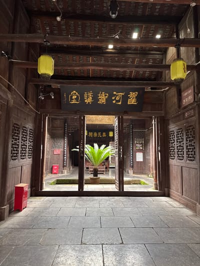 The image depicts an interior courtyard of a traditional wooden building in Zhuoshui, China. The space features wooden pillars and beams, with paper lanterns hanging from the ceiling. Inscriptions in Chinese characters adorn a plaque on the wall. Through the courtyard's openings, other buildings and greenery are visible. The floor is paved with stone slabs. There is a red fire extinguisher box on the wall.