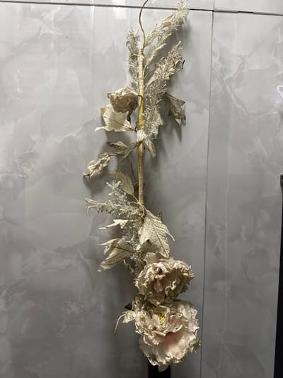 The image shows a close-up, vertical shot of a dried floral arrangement against a grey, marbled tile wall. The arrangement is a stem with dried, pale leaves and two large, pale pink and white flowers with ruffled petals at the bottom. The stem is thin and twisted, with some delicate, wispy white accents resembling dried grass or seed heads. The flowers appear to be artificial or very old, with a slightly faded and delicate appearance. The wall behind is made of large, rectangular tiles with a subtle grey marbling pattern. The lighting is soft, casting minimal shadows. There are no people or animals present. The environment suggests an indoor setting, possibly a home or a decorative display area. The context of Sumgayit, Azerbaijan, is not visually indicated within the image itself.