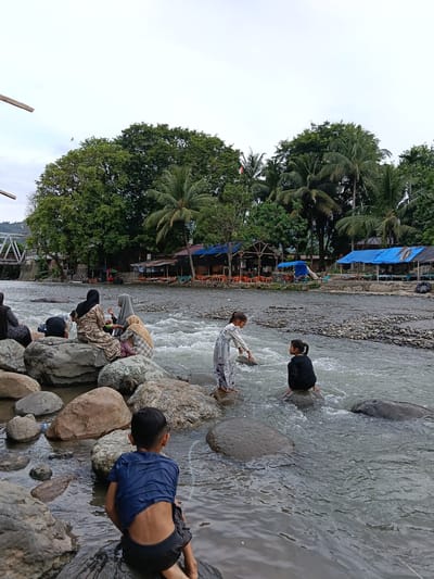 The image captures a scene at a riverbank, possibly in Bateiiliek, Indonesia, where a group of people are enjoying the water. In the foreground, a young boy wearing a blue t-shirt and dark shorts sits on a large, wet rock, facing away from the camera and towards the river. The river flows with moderate current over a rocky bed.
Several individuals, primarily women and children, are positioned on rocks closer to the bank. Three women are seated together on large rocks, two of whom are wearing headscarves. One woman is holding a small bowl and a spoon, possibly eating or offering food. Another woman is partially visible on the far right, wearing a patterned garment.
Further into the river, two young girls are interacting with the water. One girl, in a light-colored patterned dress, stands on a rock and appears to be reaching towards the water. The second girl, wearing a black outfit, sits on a rock in the river.
The background features a lush green environment with numerous trees, including palm trees. Along the opposite bank, there are several structures with blue and dark-colored roofs, some with orange chairs and tables set up, suggesting a recreational area or food stalls. A metal bridge with a white railing is visible in the distance to the left, crossing the river. The sky is overcast, indicating a cloudy day. The overall atmosphere appears relaxed and familial.