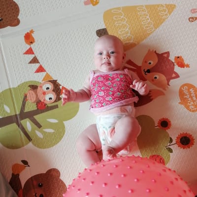 A baby is lying on its back on a playmat. The baby is wearing a pink top with strawberries and a diaper. The playmat has cartoon animals and patterns. The baby's legs are slightly bent, and its arms are outstretched. The image appears to be taken from a slightly elevated angle. There is a large pink spiked ball in the foreground, partially obscuring the baby's lower body. The background is blurred, suggesting it is a domestic indoor environment. There are no visible indications of Trubacheyevka, Russia, in the image itself.