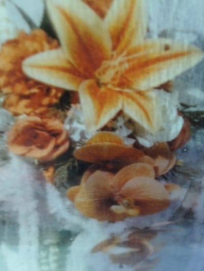 The image displays a close-up of an arrangement of flowers. The primary focus is a large, star-shaped lily with prominent orange and yellow stripes. To the left and below the lily are several smaller, peach-colored roses, some appearing more open than others. The background is a soft, light blue and white, suggesting a fabric or textured surface, possibly a tablecloth or a decorative filler. There are no people or discernible elements of an urban environment, such as buildings or landmarks, in the image. The floral arrangement appears to be indoors or in a setting where natural outdoor elements are not visible. The location of Cottrueng, Indonesia, is not evident from the visual information presented in the image.