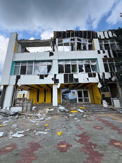 A forensic view of a three-story building, severely damaged, located in Staryi Saltiv, Ukraine, under a partly cloudy sky. The structure, featuring a modern design with white, yellow, and black facade elements, shows extensive blast damage. The uppermost story exhibits significant structural failure, with a substantial portion of its roof and outer wall destroyed, exposing internal concrete and rebar. Windows across all three visible levels are shattered, with missing panes and damaged frames. White and black facade panels are torn, detached, and partially hanging, exposing underlying structure or insulation. The ground floor entrance, characterized by yellow architectural elements, has a set of steps leading to shattered glass doors. Shattered glass, white and black facade fragments, and other construction debris are strewn across these entrance steps and the adjacent paved walkway, which features interlocking red and grey pavers. A dark green tree is partially visible on the right side of the building. No individuals are present in the scene.