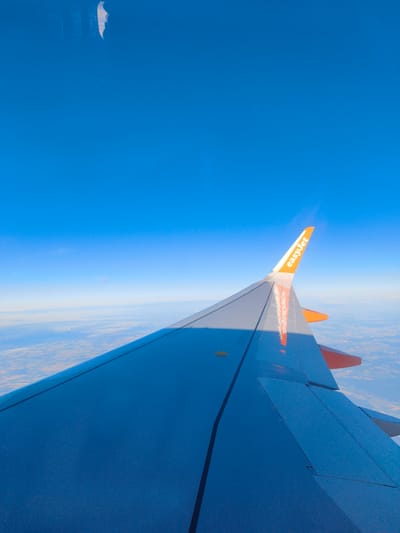 The image is an aerial view from an airplane, capturing the wing and a vast expanse of blue sky. The wing, painted in shades of blue and grey with orange accents, extends from the right side of the frame. The EasyJet logo is visible in orange on the wingtip. Below the wing, a landscape of fields and what appear to be small structures stretches towards the horizon, softened by atmospheric haze. The location in Schipkau, Germany is not directly discernible from the visual information presented in the image.