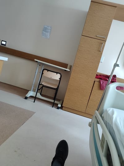 The image depicts a hospital room in İnegöl, Turkey. In the foreground, a person's leg and foot wearing a black shoe are visible, angled as if they are standing or sitting. To the right, a hospital bed with white bedding and a blue stripe is partially shown, with an IV pole next to it. In the background, a tall wooden cabinet with horizontal slat detailing and silver handles is present. To the left of the cabinet, a foldable chair with a light-colored seat and backrest is positioned under a height-adjustable desk with a white tabletop and a grey metal frame. The wall above the desk features a mounted digital display and a document. The floor is a light-colored linoleum with sections of a different textured flooring. A red garment is draped over a surface to the right of the cabinet. The overall environment suggests a clean and functional hospital setting.