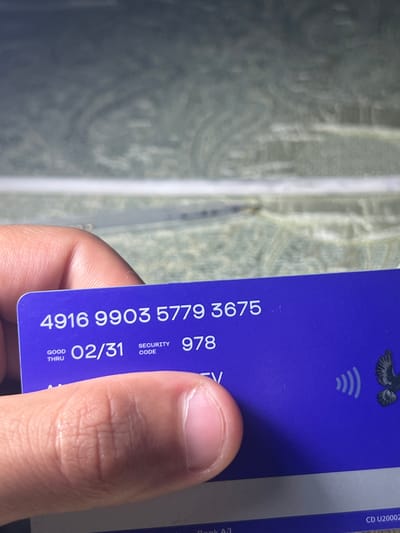 The image shows a close-up of a purple card being held by a person's thumb and index finger. The card displays alphanumeric text and a graphic of a bird with outstretched wings, as well as a contactless payment symbol. The visible text includes "4916 9903 5779 3675", "GOOD THRU 02/31", "SECURITY CODE 978", and "TV". The background of the image is out of focus and appears to be a textured surface with a pattern of swirling lines, possibly resembling wood grain or marble. The lighting suggests the image was taken indoors or in an area with ambient light. The location is identified as Romitan, Uzbekistan.