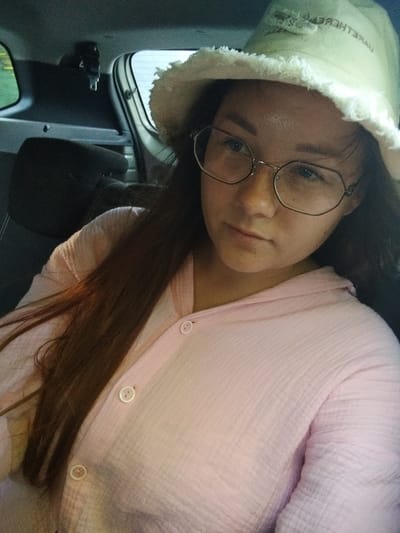 The media file is a close-up, first-person perspective selfie taken inside a vehicle, likely a car or van. The subject is a young person wearing glasses and a light-colored bucket hat with a frayed brim. The hat has some faint, unreadable text on it. The person has long, dark brown hair and is wearing a light pink, button-up shirt made of a textured fabric, possibly gauze or muslin. The shirt has two visible buttons.
The background suggests the interior of a vehicle. To the left, a green tinted window is visible, possibly showing an outdoor scene. The back seat headrest and part of the car's interior trim are also visible, in dark gray and black tones. The lighting suggests it could be daytime, possibly late afternoon or early evening, given the somewhat dim interior lighting. There are no strong indicators of weather conditions. The subject's expression is neutral, with a slight gaze towards the camera. The overall impression is a casual, candid moment captured while traveling. The location context is Grabovo, Russia.