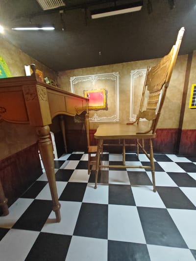 The image depicts an indoor scene within a room designed to create optical illusions, possibly a museum exhibit or attraction. The primary subjects are a large, ornate wooden chair and a wooden table. The chair is tall and intricately carved, with a high, decorative back. The table is also wooden and appears to be antique, with carved legs. A small, child-sized chair is visible beneath the table.
The setting is characterized by a dramatic black and white checkered floor that creates a strong visual effect, enhancing the illusionary nature of the room. The walls are decorated with patterned wallpaper and framed artwork, including a prominent framed picture with a red center. The lighting appears to be artificial, with ceiling lights visible.
The scene appears staged, with the furniture placed in a way that plays with perspective. There is no activity or event taking place; the image captures a static view of the room. The mood is one of visual intrigue and perhaps disorientation due to the optical illusions. There are no visible people, and no text is discernible in the image. The location context is Gongshu District, China, suggesting this is a specific attraction within that area. The overall impression is one of a surreal and engaging environment designed to trick the eye.