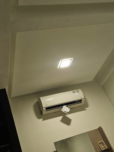 The image depicts an interior view of a room in Padajaya, Indonesia. An off-white ceiling features a centrally illuminated square LED panel light. Below, a white Midea brand split-unit air conditioner is mounted on an off-white wall, displaying "23" on its digital screen. A small white rectangular packet is affixed to the front panel of the air conditioner, casting a shadow on the wall below it. A portion of a rectangular mirror is visible in the lower right, reflecting a dark area and a small framed picture. No individuals are present in the scene.