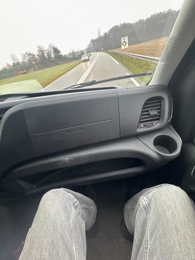The scene is observed from the interior passenger seat of a vehicle traveling on a wet two-lane road in the vicinity of Haguenau, France, under an overcast sky. The lower portion of the image displays the passenger's legs, dressed in light grey denim jeans and dark footwear, positioned within the footwell. The vehicle's dark dashboard occupies the mid-ground, featuring a passenger airbag panel inscribed with "AIRBAG," a right-side air vent, and an integrated circular cup holder. Through the windshield, the asphalt road, slick with moisture, extends forward and curves gently to the right. A silver-colored sedan with activated headlights is visible ahead on the road. Roadside features include grassy embankments and a metal crash barrier on the right. A prominent white rectangular sign on the right verge indicates "Haguenau" and an upcoming roundabout, with "PRE-SIGNALISATION PROCHAINE SORTIE" printed above the roundabout symbol. The distant landscape comprises dense trees and foliage beneath a grey, cloudy sky.