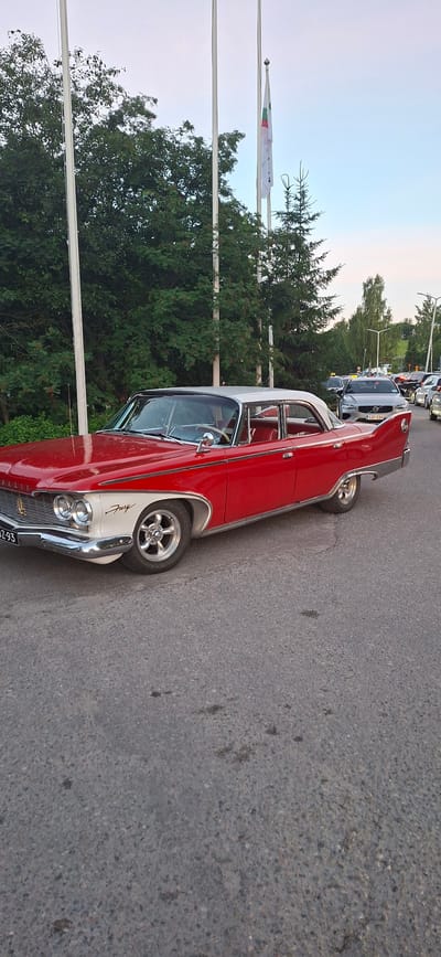 The image depicts a classic red and white two-door sedan, identified as a 1957 Plymouth Fury, parked on an asphalt road. The car is the primary subject, positioned slightly to the left of the frame, facing right. Its distinctive chrome grille, dual headlights, and iconic tail fins are visible. A license plate with the characters "SZ-93" is affixed to the front. The car's interior, featuring red seats, is partially visible through the windows.
In the background, several other vehicles are present, including a silver Volvo sedan and what appears to be a taxi. Trees and flagpoles line the right side of the road. One flagpole displays a flag with green, white, and red vertical stripes and a symbol. The environment suggests an outdoor setting in a town or city, possibly during daylight hours with overcast skies. The image was taken in the city of Salpakangas, Finland.