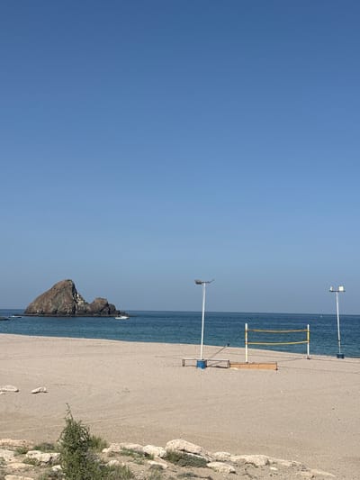 The image depicts a beach scene in Al Aqah, United Arab Emirates. The photograph is taken horizontally, with a clear blue sky occupying a large portion of the upper left quadrant. The right side of the image features a sandy beach that extends from the upper right corner downwards. Adjacent to the beach is the ocean, which forms a border between the sand and the sky. Several beach volleyball nets and poles are erected along the shore. In the distance, a rocky outcrop rises from the sea. Vegetation and rocks border the beach on the right side of the image. There are no visible people in the scene.