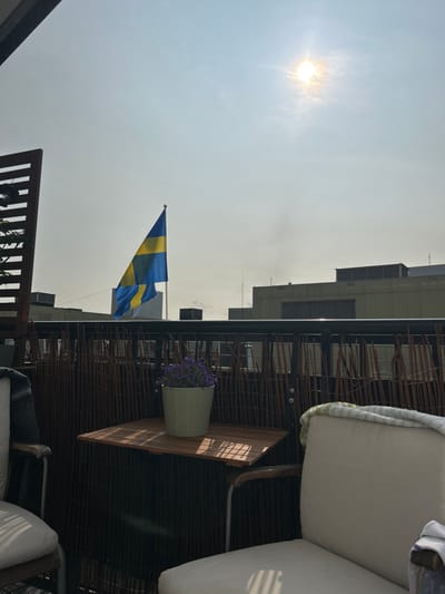 The image shows a balcony in Malmö, Sweden, located at coordinates 55.594 latitude and 13.01 longitude. The balcony features a black metal railing with a woven brown wicker fence paneling behind it. A small wooden table with a light wood top sits in the center of the balcony, and two chairs are placed to the left and right of it. There are two people on the balcony, but they are only partially visible; only the back of a chair and the back of a person sitting in the chair on the left can be seen, and only the chair on the right is visible. A small potted plant with purple flowers sits on the table. A Swedish flag is flying from a pole to the left of the balcony. The sky is clear and bright with the sun shining brightly in the middle of the image. The buildings in the background are out of focus, but they appear to be made of concrete and painted in a light grey color. The overall tone of the image is peaceful and relaxing.