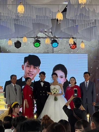 This image depicts an indoor wedding reception, likely held in Bình An, Vietnam, given the cultural context. The central focus is on the bride and groom, who are standing together, smiling. The bride is adorned in a white, strapless wedding gown, holding a bouquet of white roses and green foliage. The groom is dressed in a black tuxedo with a bow tie.

Behind the couple, a large LED screen displays close-up images of their smiling faces. Surrounding them are several guests and family members. A woman wearing a traditional red Vietnamese Ao Dai is visible, along with a man in a grey suit and a dark tie, and another man in a dark blue suit with a striped tie. A person in a black shirt, possibly a photographer or event staff, is holding a remote device. In the foreground, a young girl with a yellow headband is partially visible among the attendees.

The venue is elaborately decorated, featuring numerous white, layered fabric installations that resemble abstract clouds or petals suspended from the ceiling. These decorations are interspersed with delicate string lights. Decorative light fixtures shaped like glowing yellow tulips or lotus flowers are also prominent. Modern stage lights, emitting vibrant red, green, and blue hues, are positioned around the area, creating a dynamic and festive ambiance. The event is characterized by a celebratory and joyous atmosphere, as indicated by the smiles of the main subjects. No explicit text is visible in the image.
